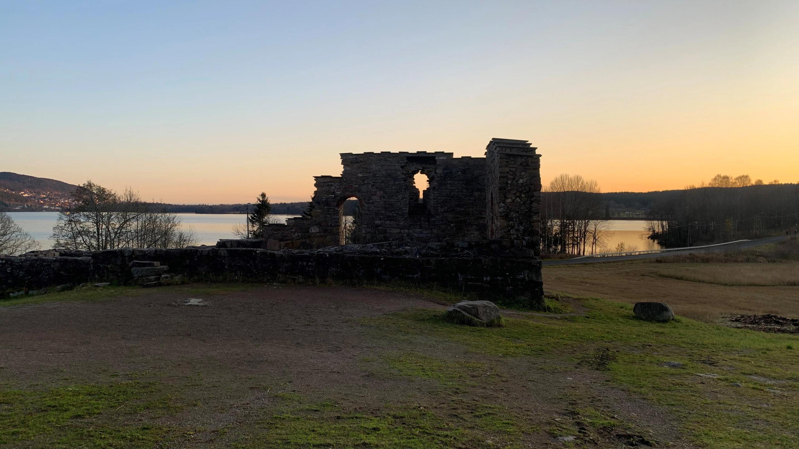 Ruins of a stone structure overlooking a peaceful lake at sunset, surrounded by grasslands and distant hills.