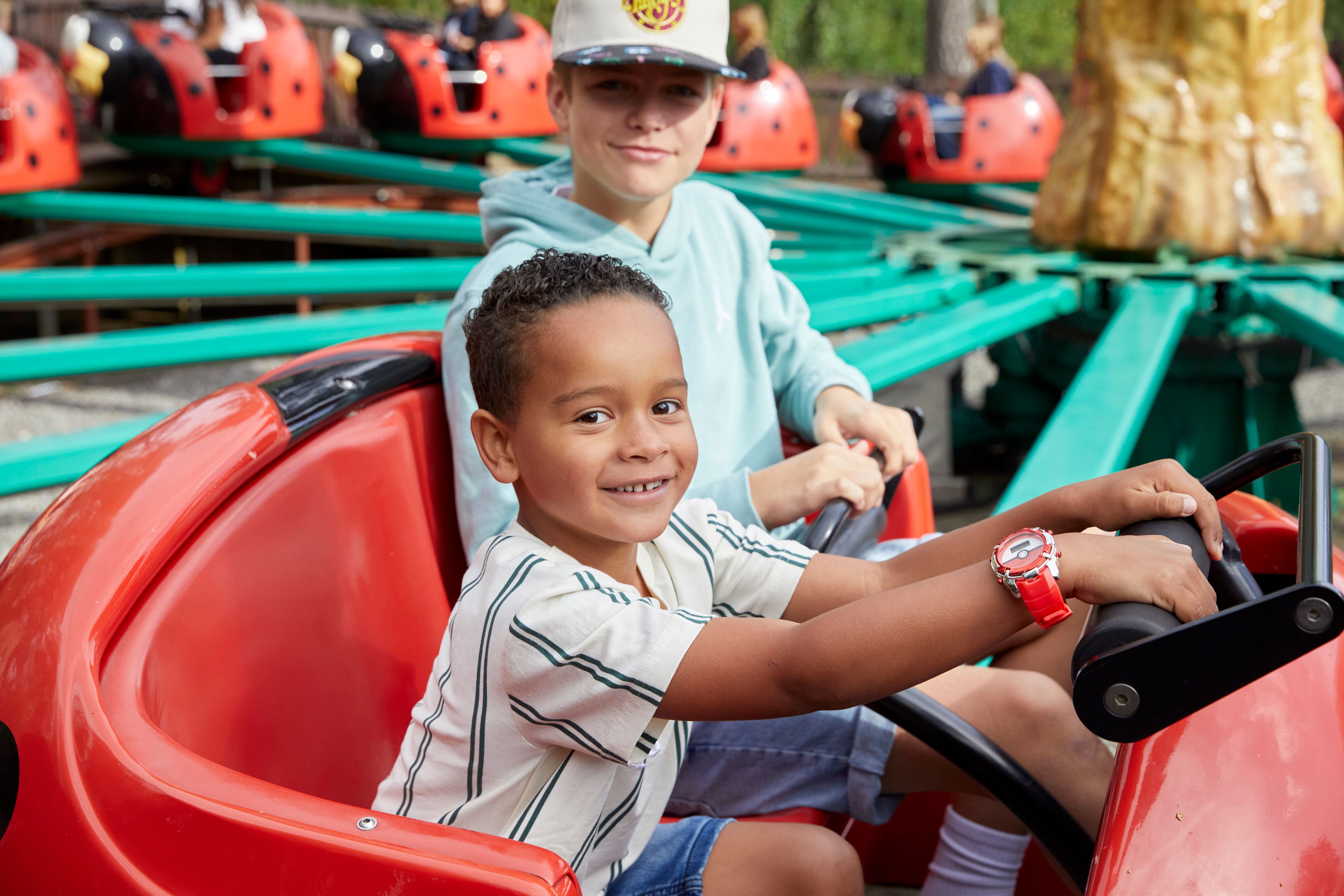 A child sits in the front seat of a red ladybug ride vehicle, holding the steering wheel, while an older child sits behind.
