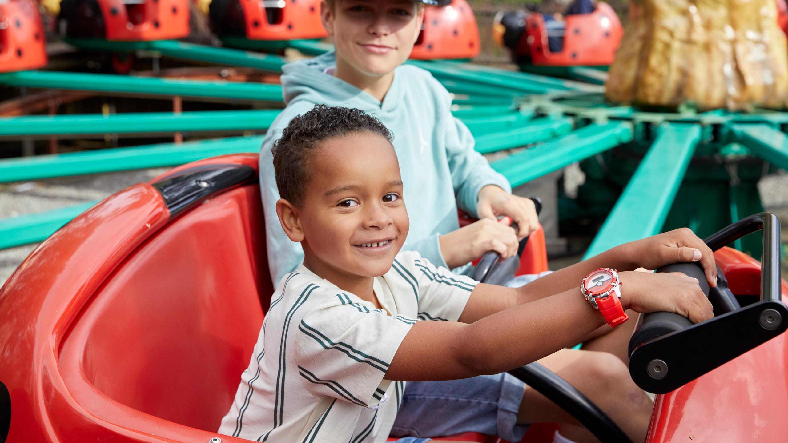A child sits in the front seat of a red ladybug ride vehicle, holding the steering wheel, while an older child sits behind.