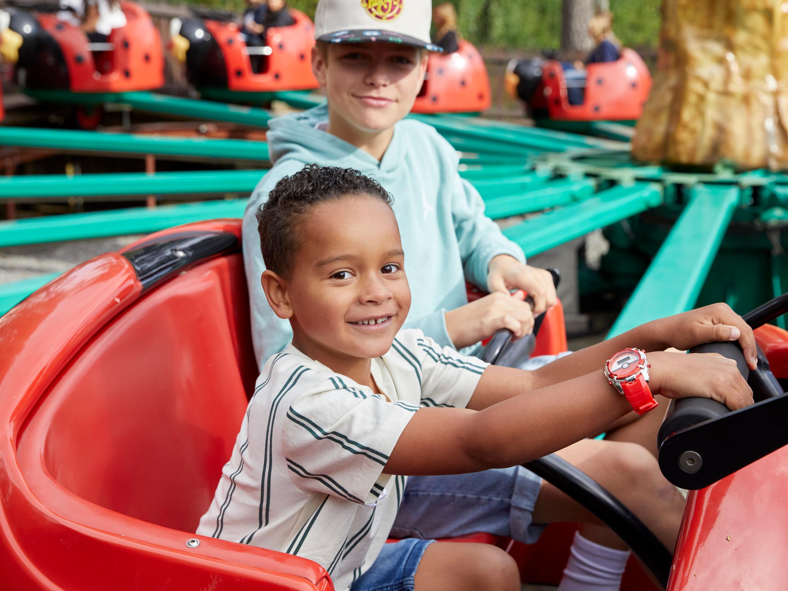 A child sits in the front seat of a red ladybug ride vehicle, holding the steering wheel, while an older child sits behind.