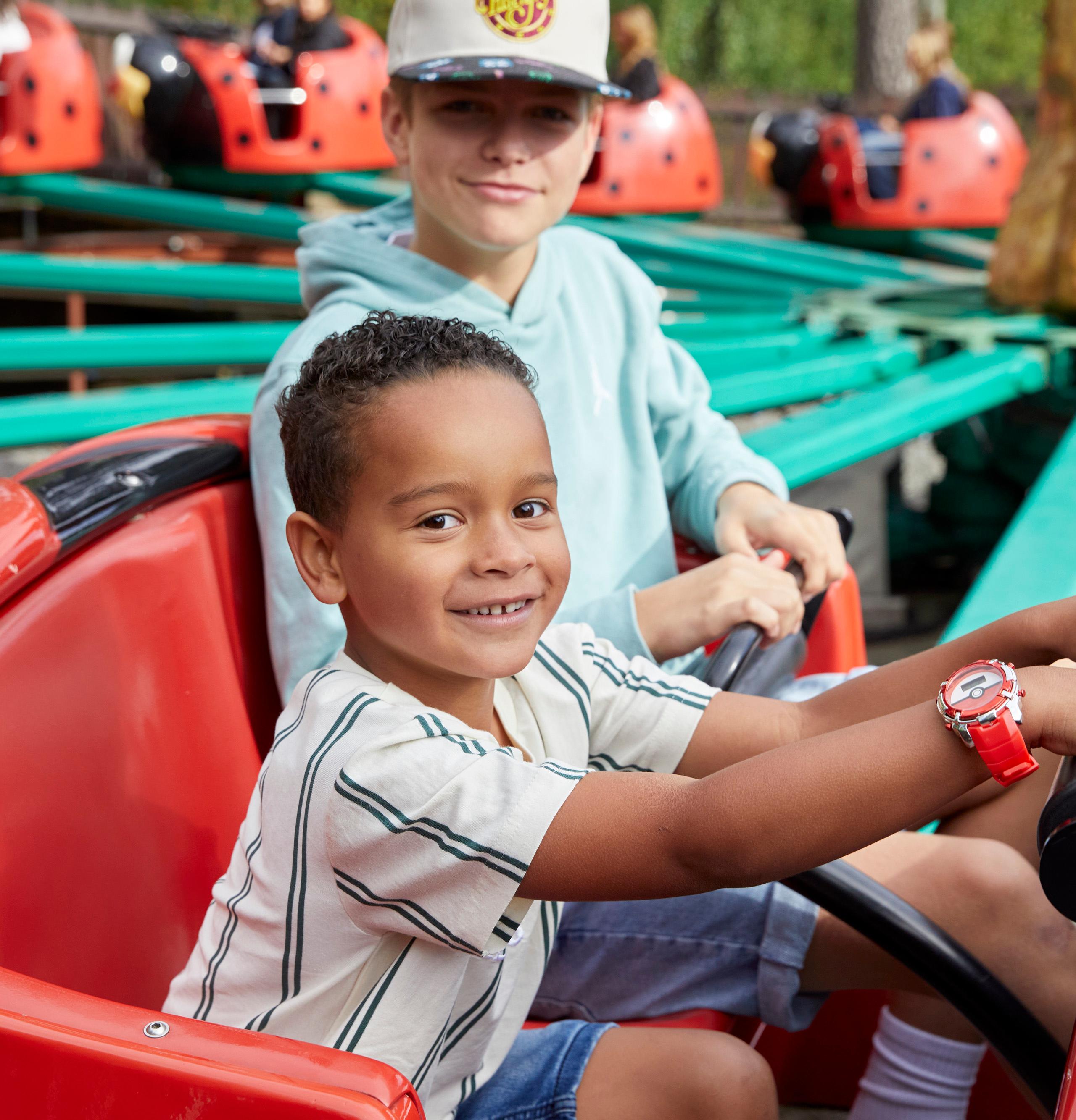 A child sits in the front seat of a red ladybug ride vehicle, holding the steering wheel, while an older child sits behind.
