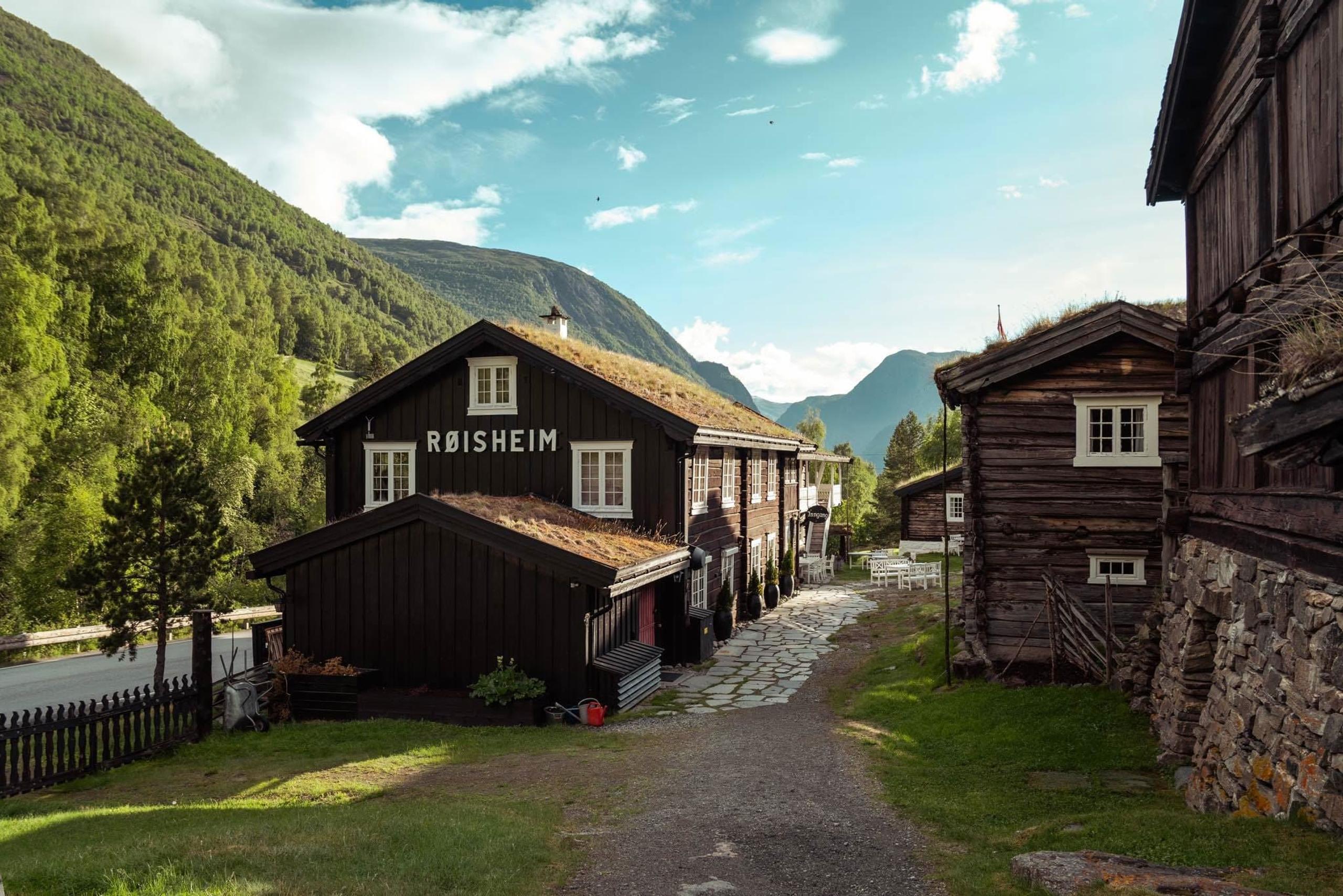 Traditional dark wooden buildings with grass roofs at Røisheim, set along a gravel path in a green mountain valley under a blue sky.