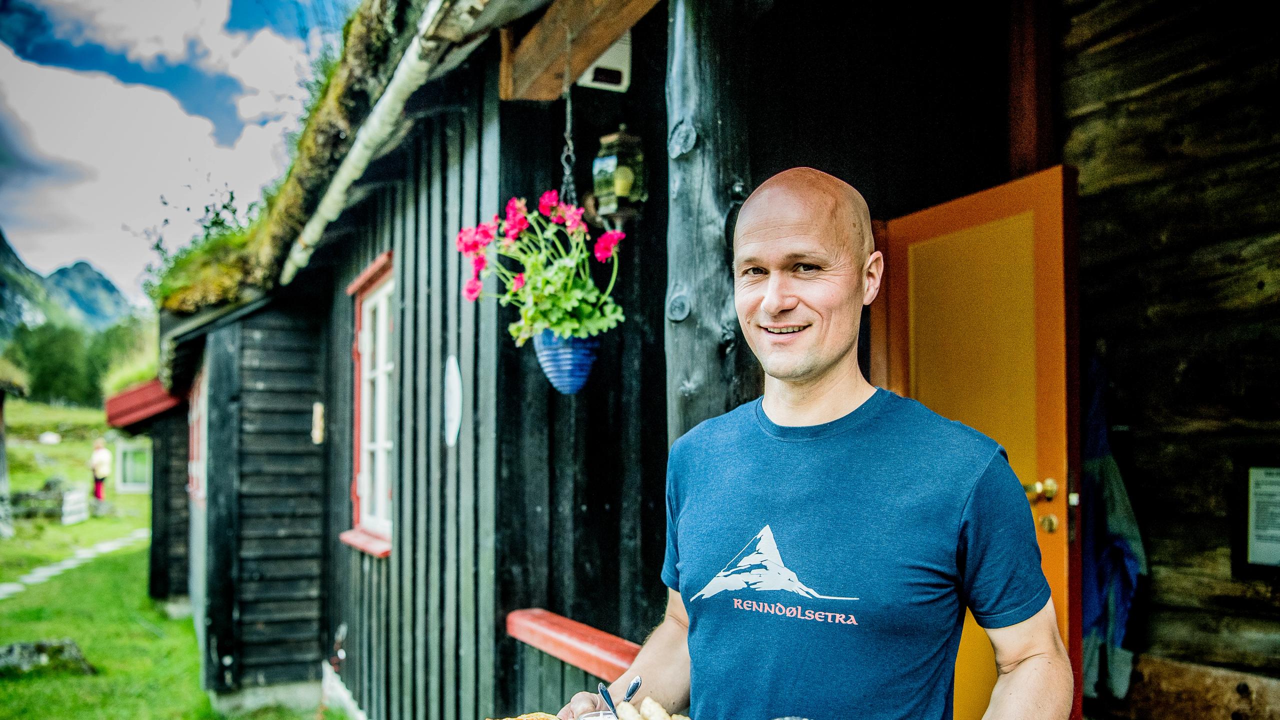 En person som jobber ved Renndølsetra holder et serveringsbrett med lokale delikatesser foran en gammel trehytte. I bakgrunnen kan du skimte en fjelltopp på venstre side.