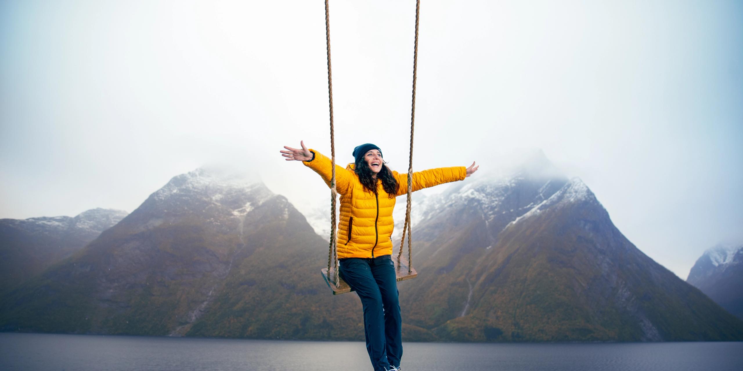 Una mujer se divierte balanceándose sobre un columpio sobre el fiordo Hjørundfjord, en la Noruega de los fiordos, con nevadas montañas al fondo.