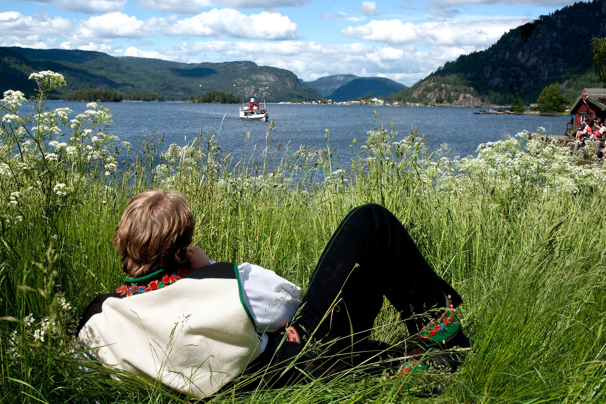 A man in the national costume Setesdalsbunad lying in the grass and watching an old steamboat on the Byglandsfjord in Setesdal, Southern Norway