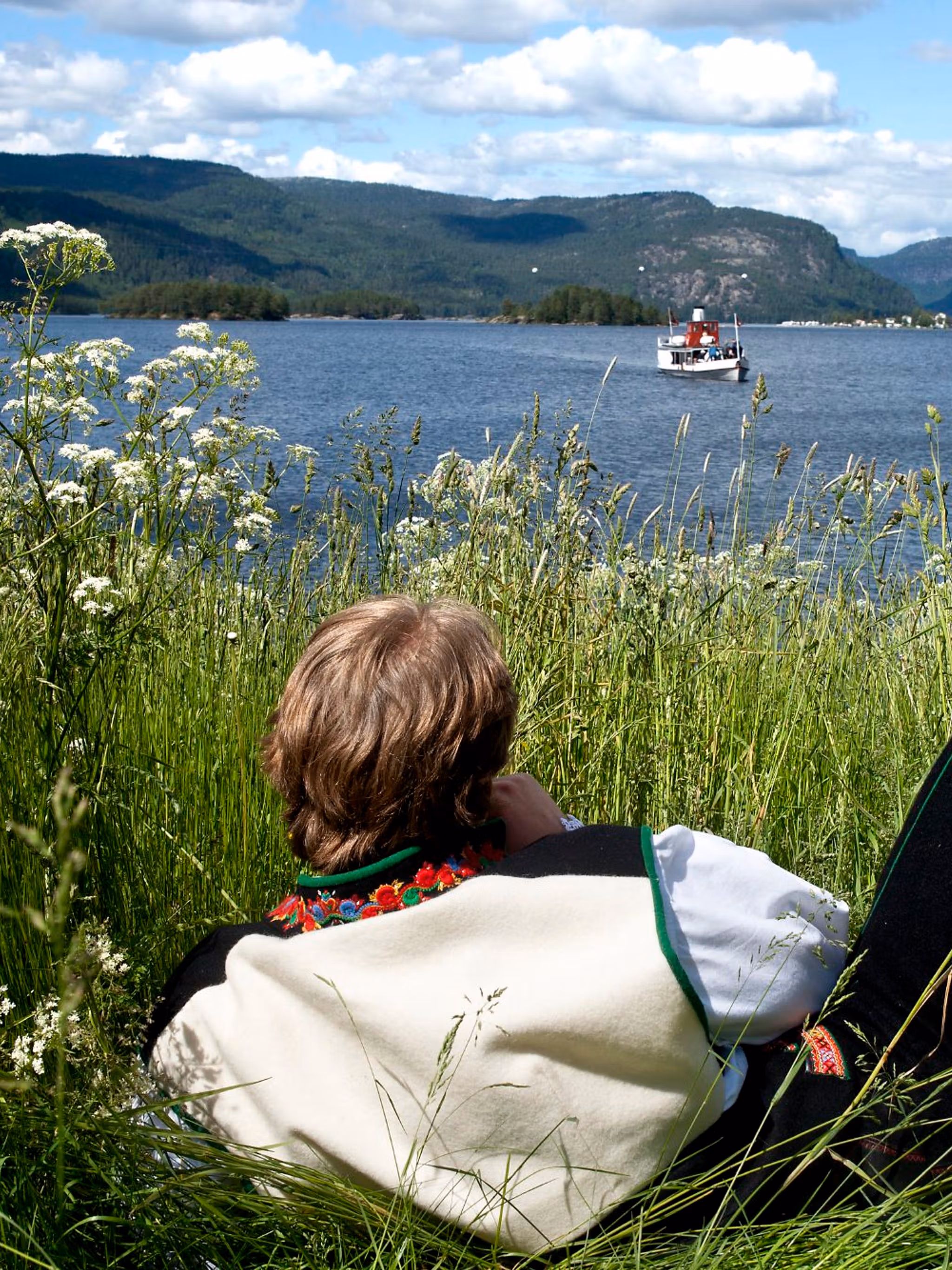 A man in the national costume Setesdalsbunad lying in the grass and watching an old steamboat on the Byglandsfjord in Setesdal, Southern Norway