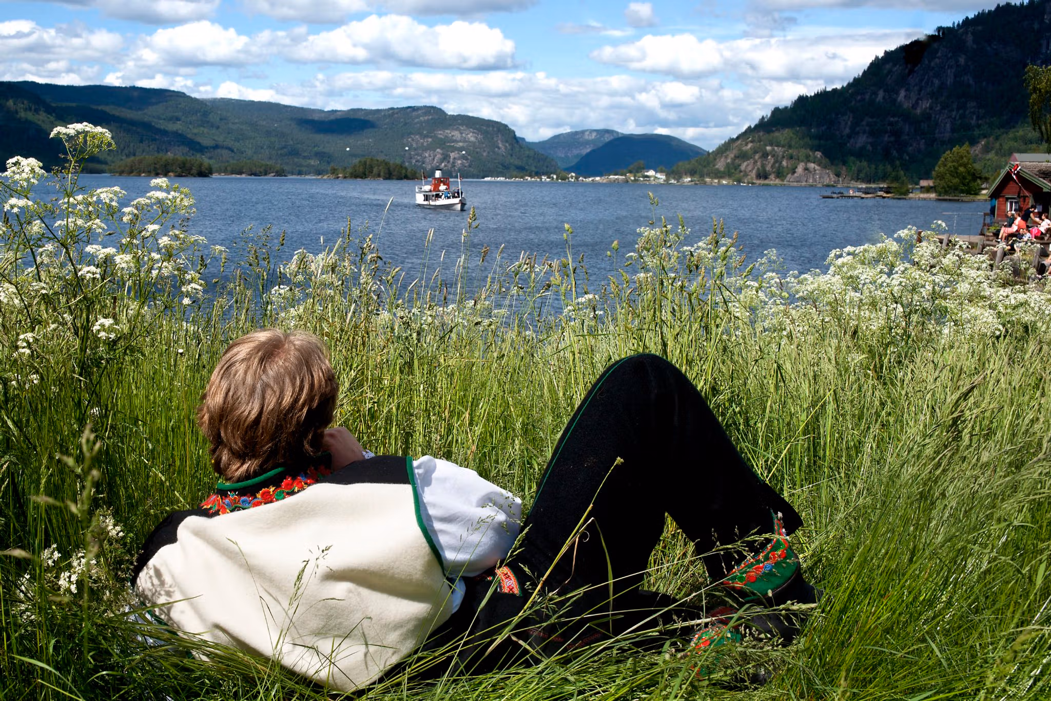 A man in the national costume Setesdalsbunad lying in the grass and watching an old steamboat on the Byglandsfjord in Setesdal, Southern Norway
