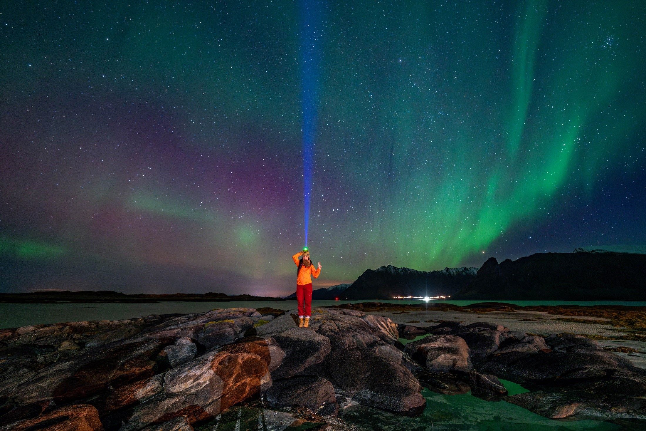A person with a headlamp stands on coastal rocks beneath a green and purple aurora borealis."