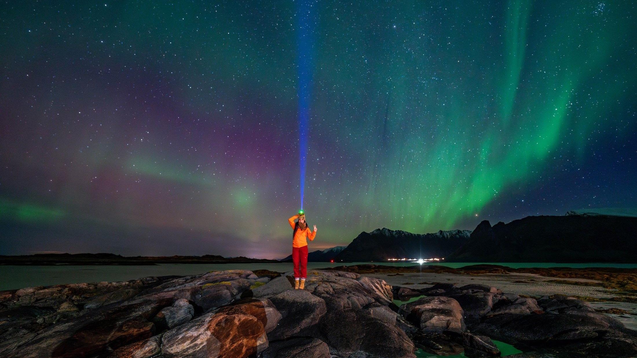 A person with a headlamp stands on coastal rocks beneath a green and purple aurora borealis."