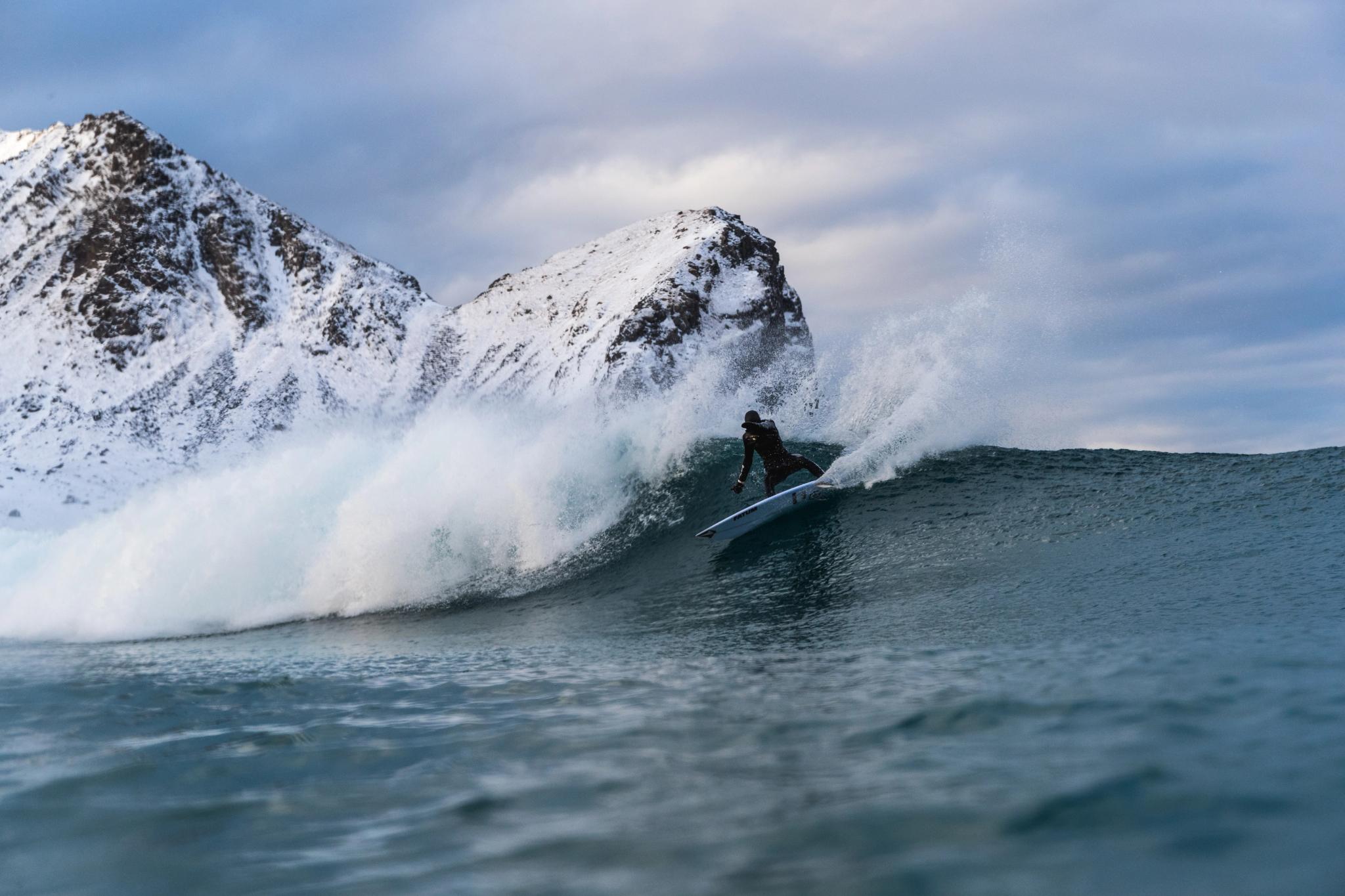 A surfer catches a breaking wave with a snow-covered mountain in the background in Lofoten in Northern Norway
