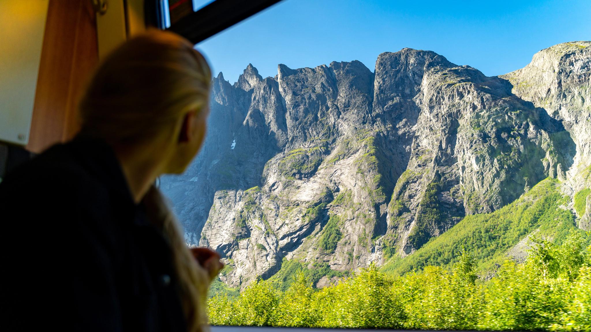 A woman looking through a train window at a towering mountain wall.