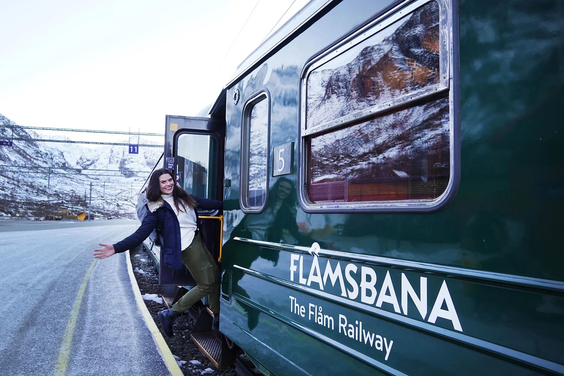 A smiling woman leans out from a green Flåm Railway train at Myrdal station, with snow-capped mountains in the background.