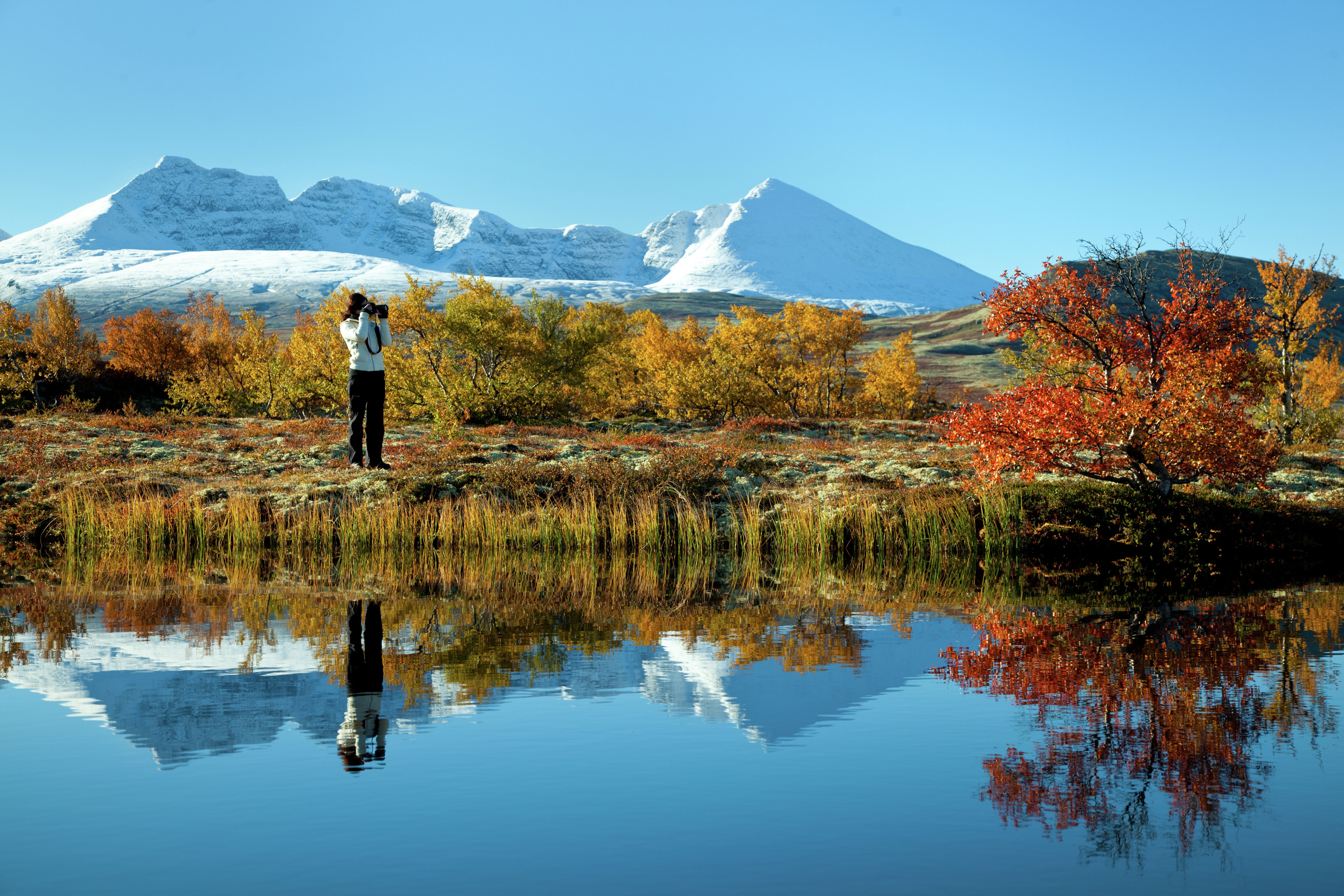 A woman taking photos of a lake and trees in autumn colours in the Rondane national park, Eastern Norway. Snowclad mountains in the background.