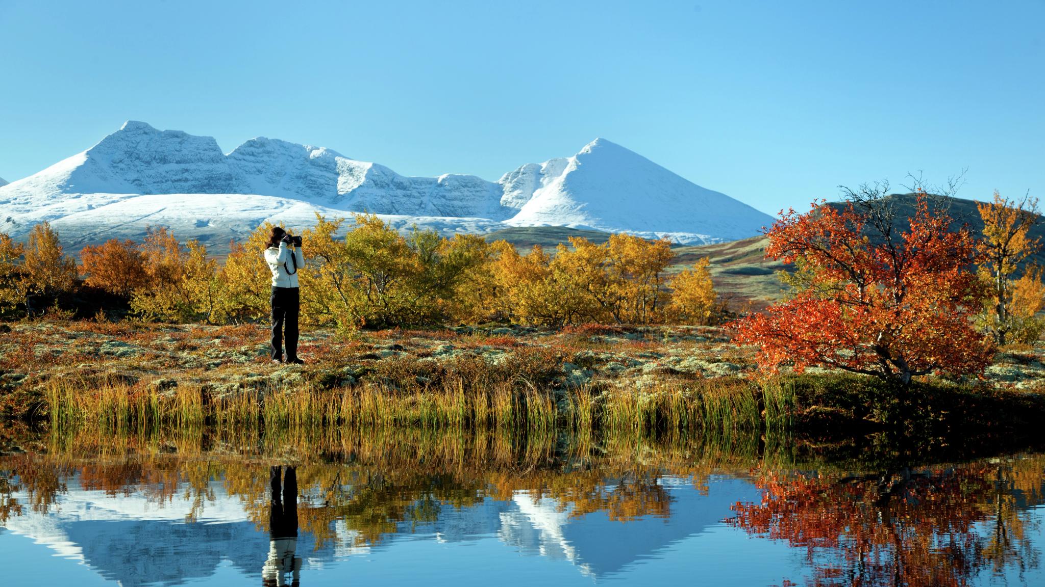Escursionista fotografa un lago e gli alberi con i colori autunnali nel parco nazionale di Rondane, Norvegia orientale. Montagne innevate sullo sfondo.