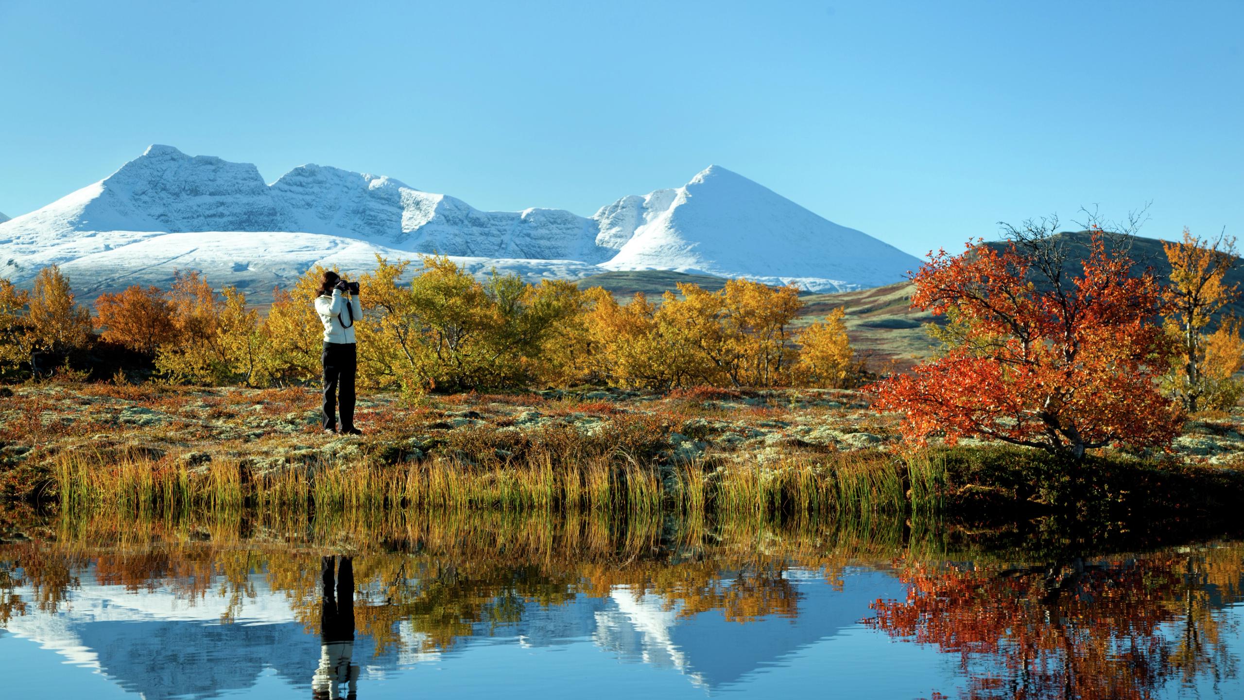 Escursionista fotografa un lago e gli alberi con i colori autunnali nel parco nazionale di Rondane, Norvegia orientale. Montagne innevate sullo sfondo.