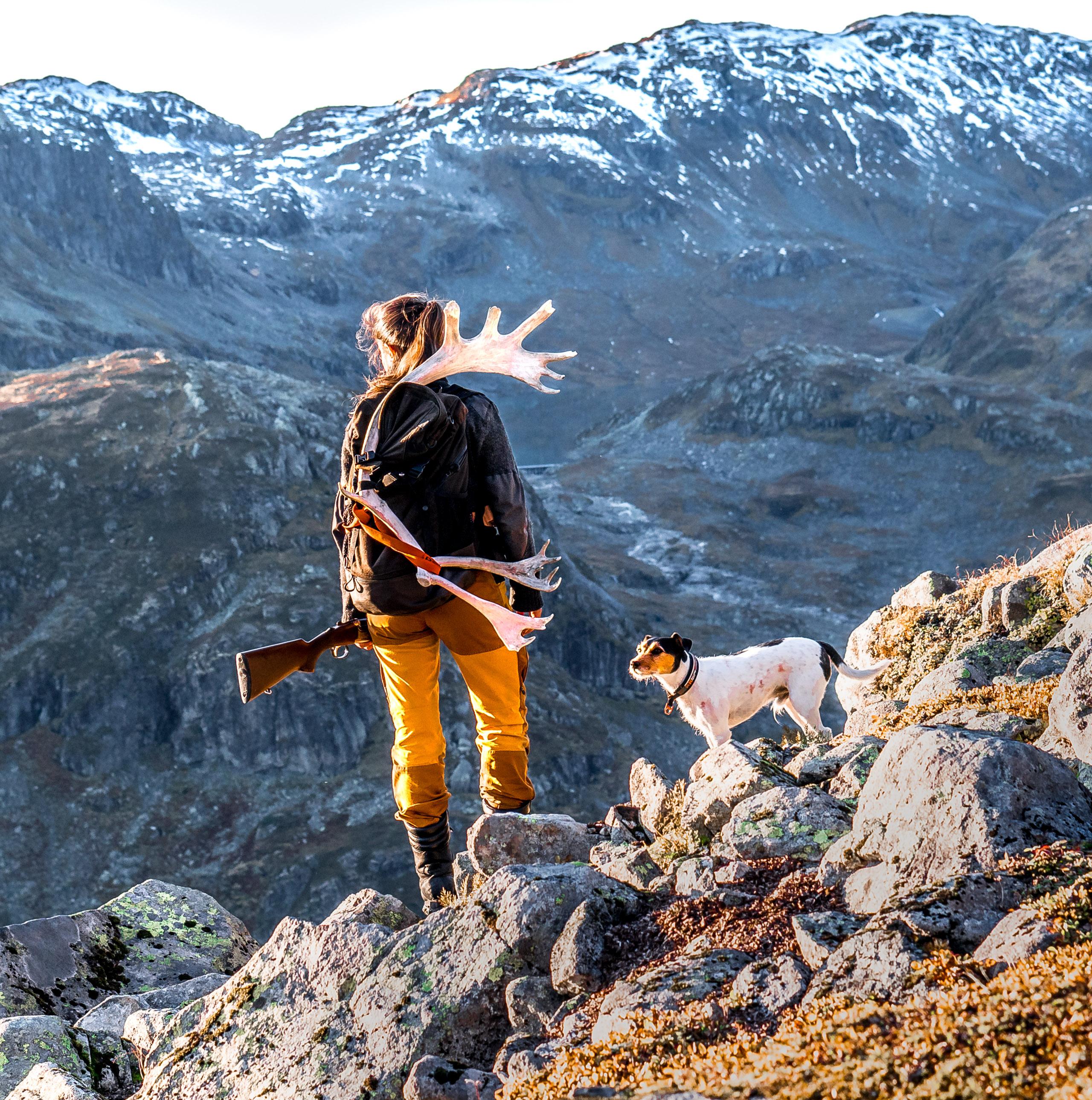 En kvinne står og ser utover fjellet med geværet i hånden og jakthunden ved sin side