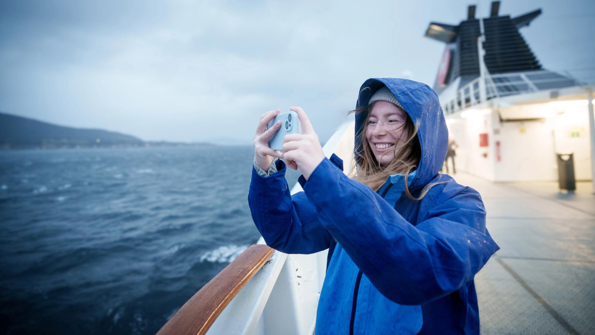 En kvinne på dekk på Hurtigruten som tar bilder i ruskevær