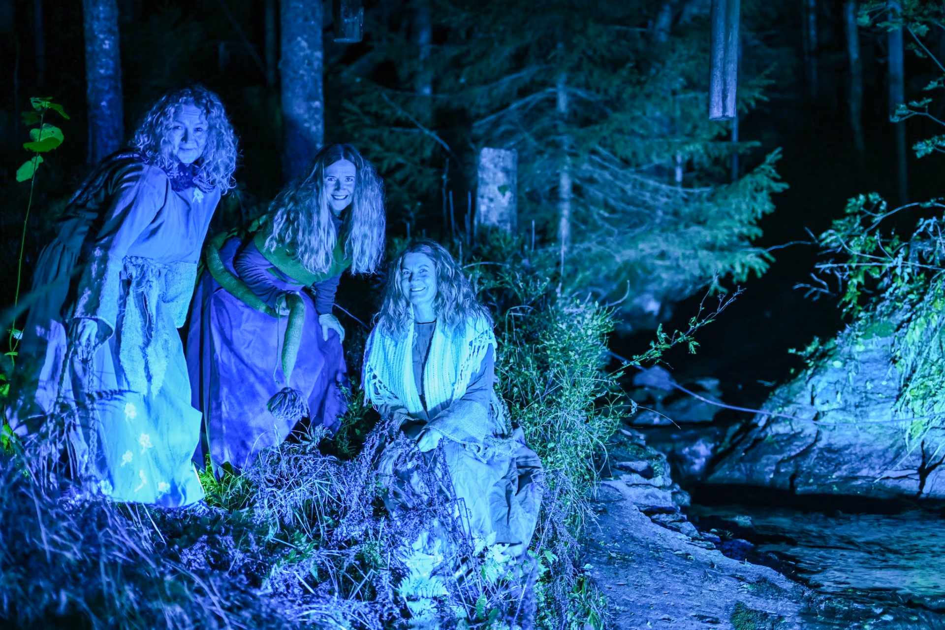 Tre kvinner er kledd i kostymer i skogen. Det er mørkt, men kvinnene er lyst opp.