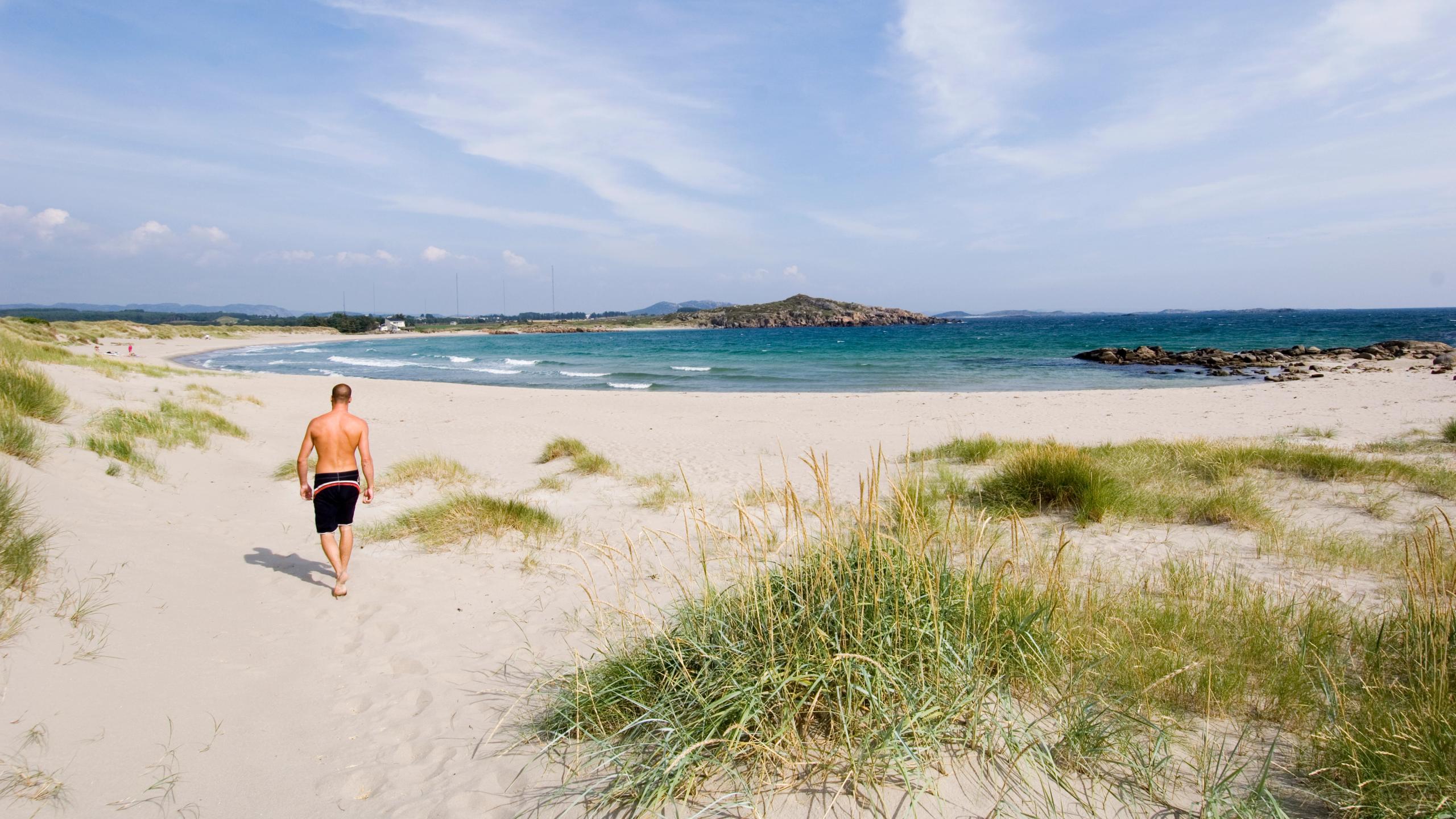 A man in a swim shorts walking on a white sandy beach in Lista, Southern Norway.