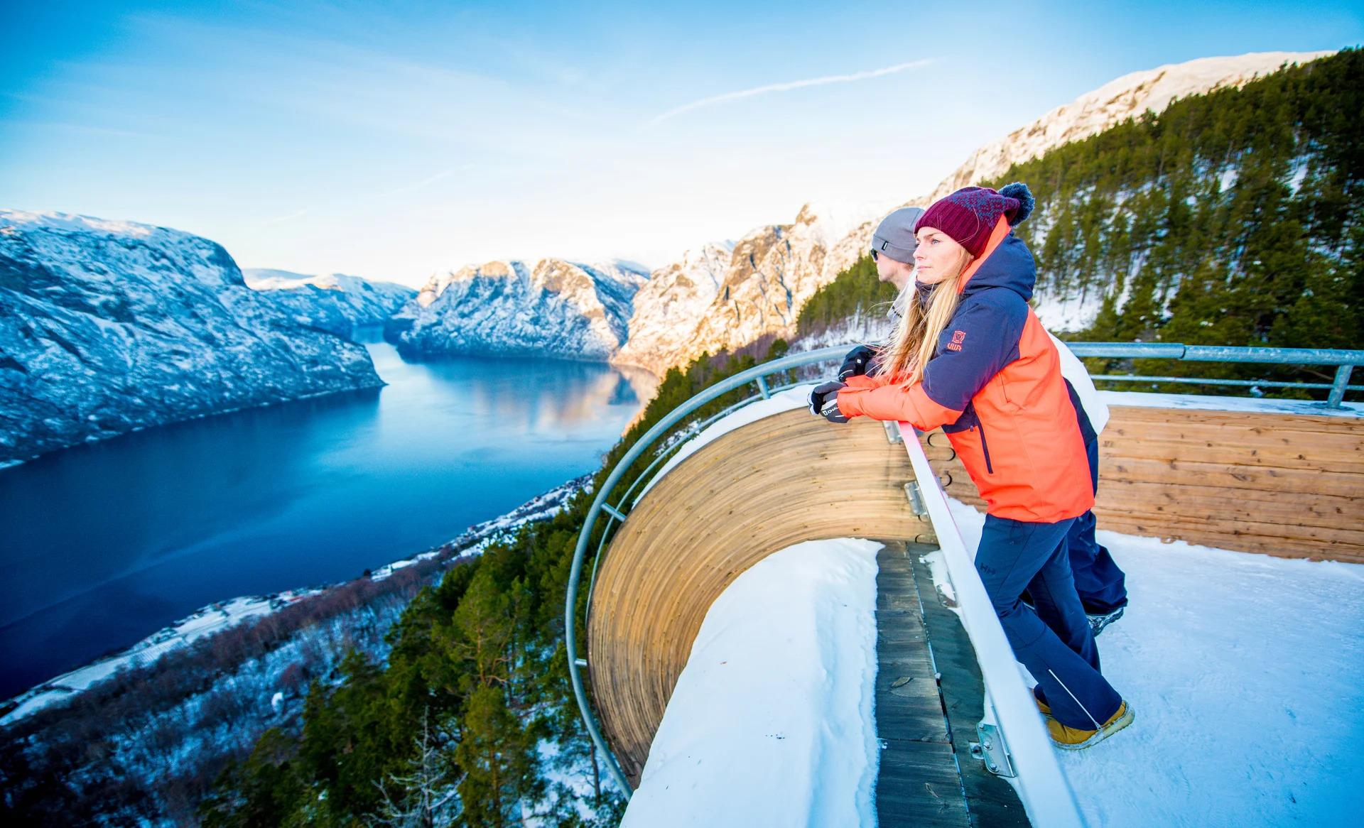 Two people in winter clothing lean on the railing at Stegastein viewpoint, a curved wood-and-steel platform overlooking the Aurlandsfjord and snow-capped mountains.