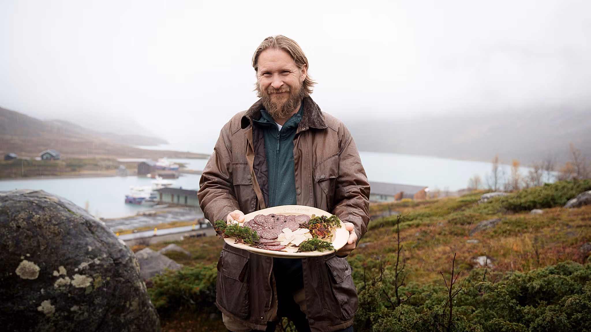 En mann i naturen holder et trefat med spekepølse.