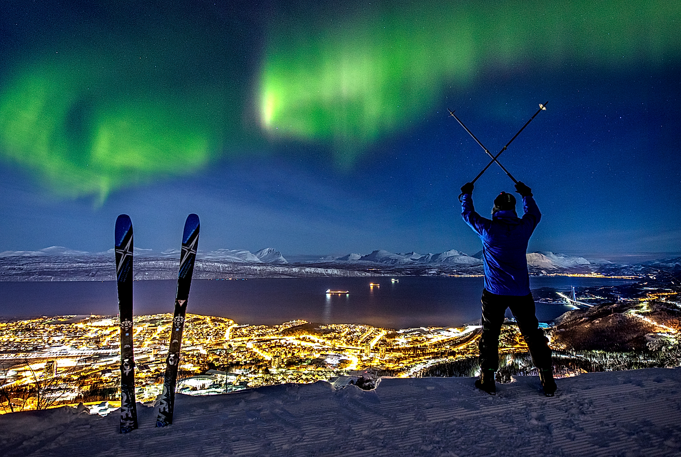 En mann kjører alpint med utsikt over Narvik by og spektakulært nordlys
