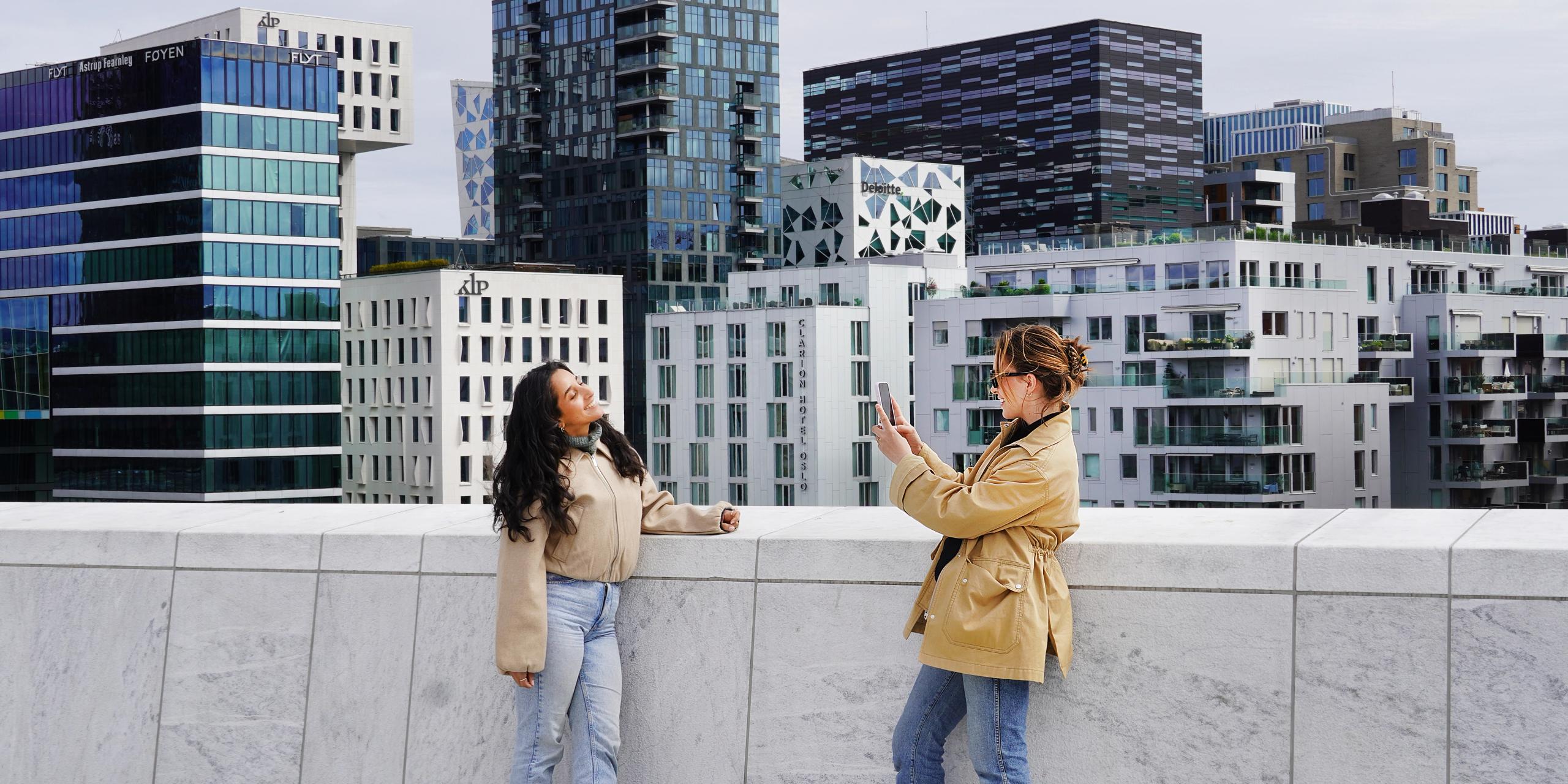Two young women standing on the rooftop of the Oslo Opera House in Norway, capturing a photo with the city skyline in the background