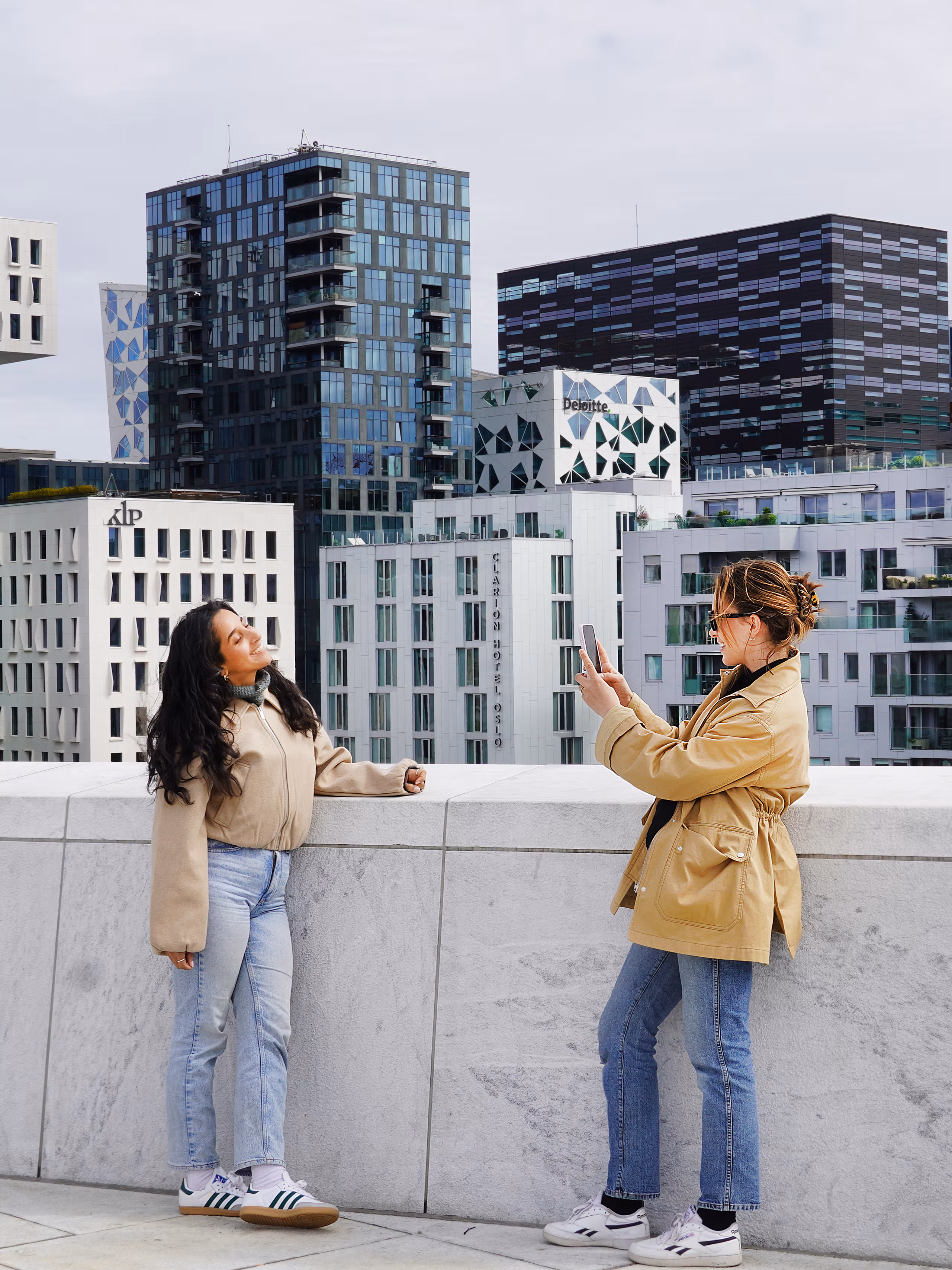 Two young women standing on the rooftop of the Oslo Opera House in Norway, capturing a photo with the city skyline in the background