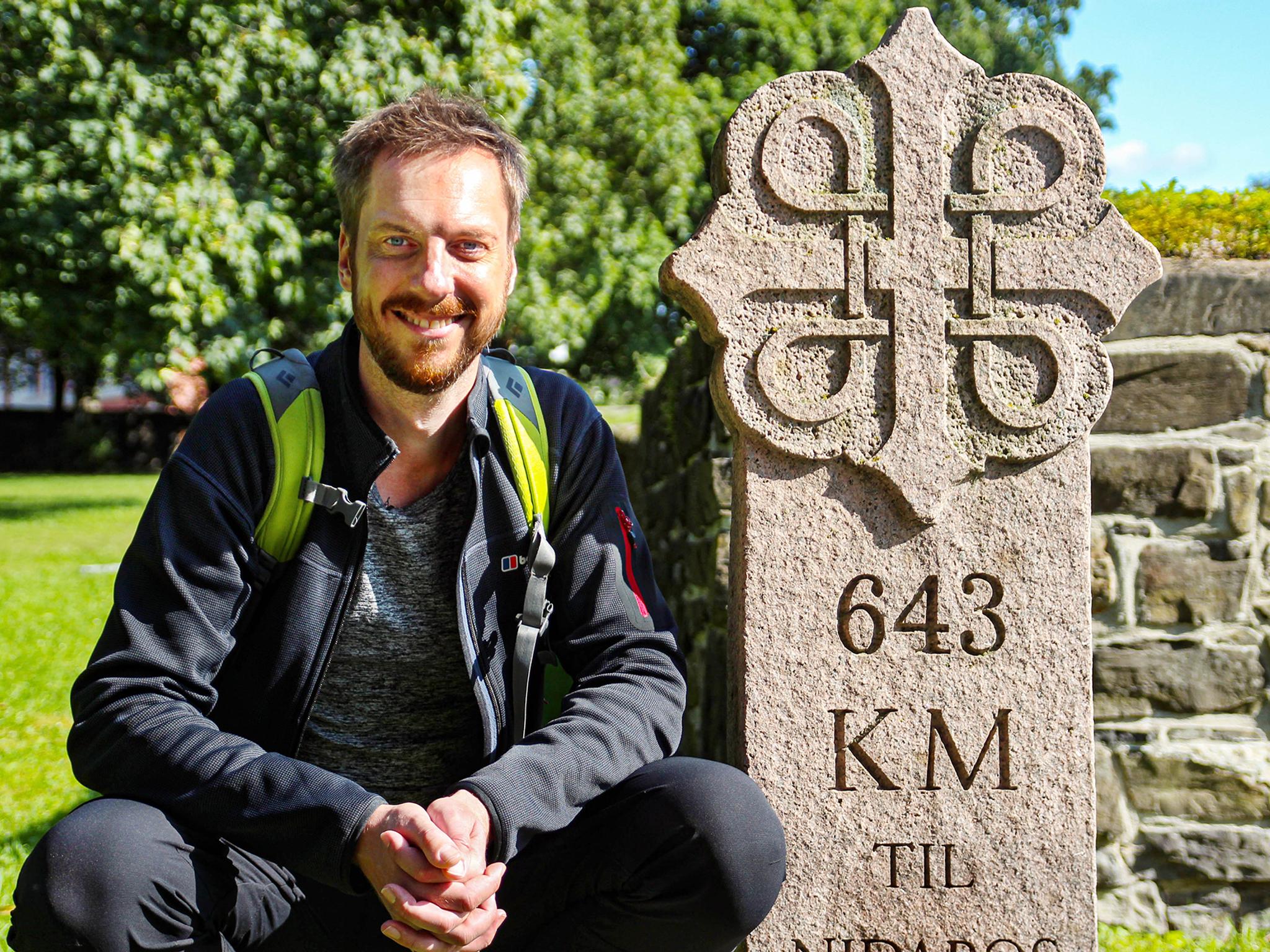 Pelgrim Kes Blans naast een bord dat de start aangeeft van de pelgrimsroute St-Olavsweg van Oslo naar Trondheim, Noorwegen.