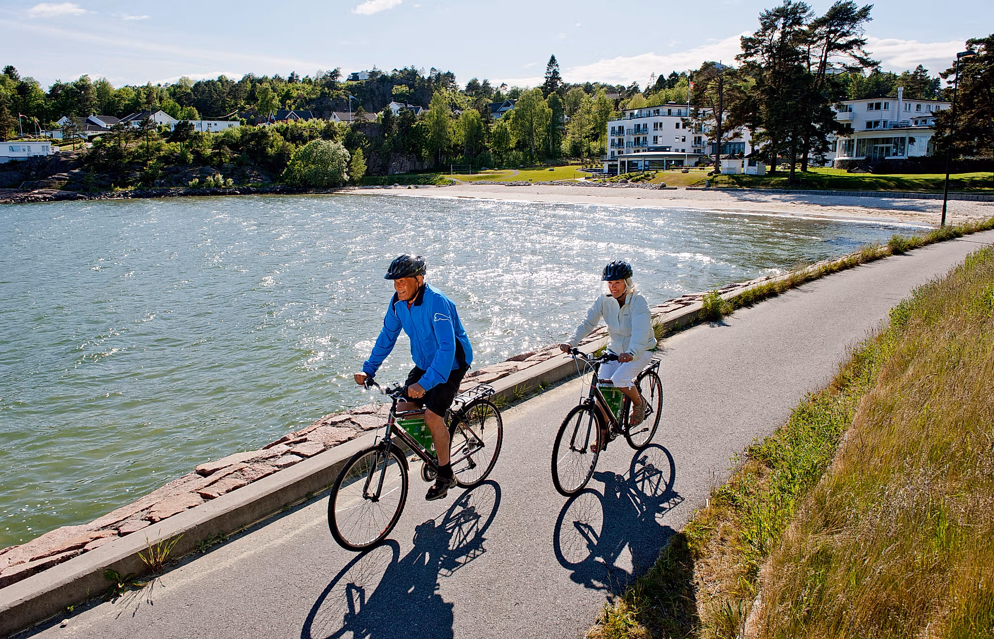 A man and a woman cycling along the sea on a sunny day in Fevik, Grimstad, Southern Norway