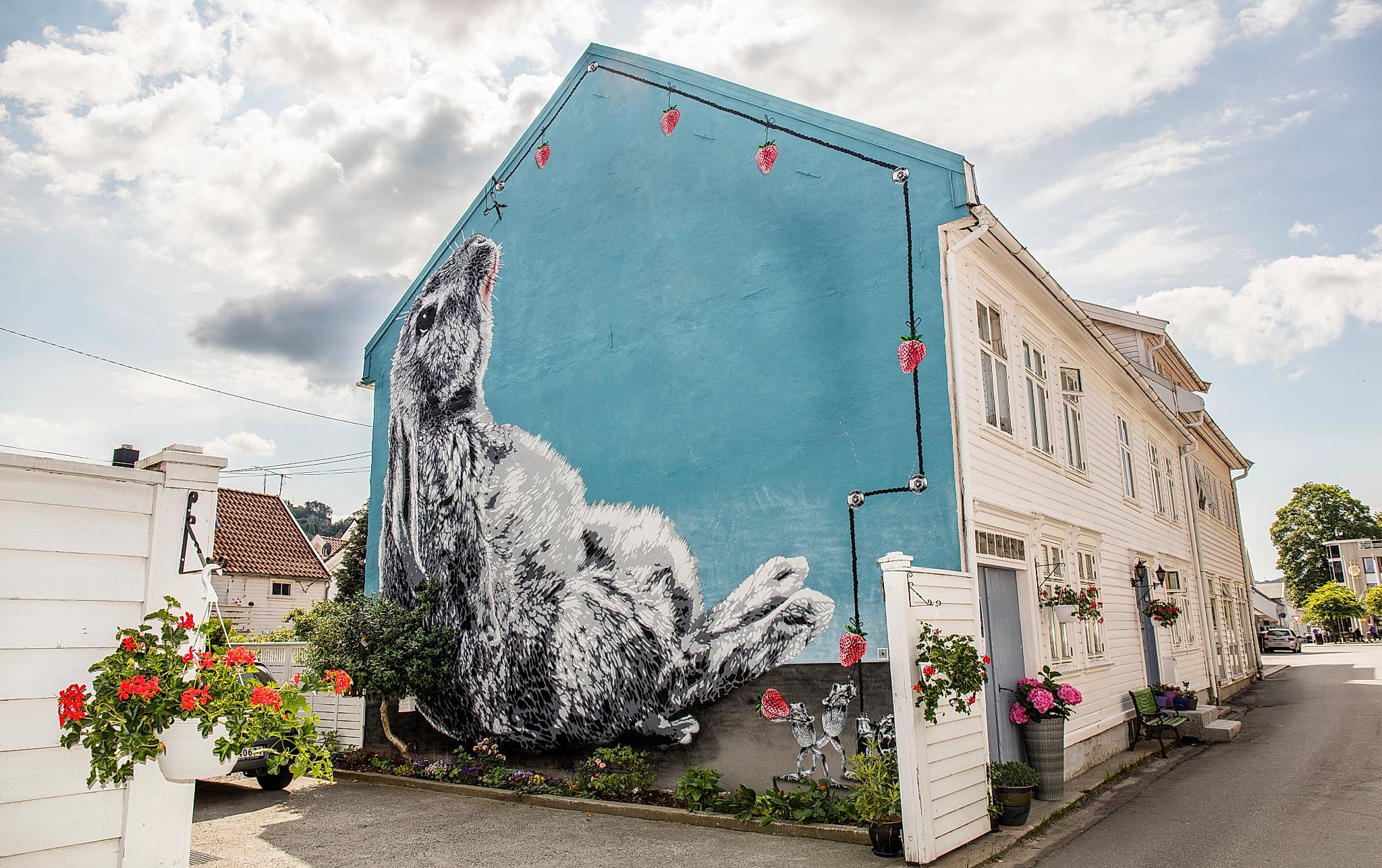 Street art covering the entirety of the short side of a white wooden house in Flekkefjord, Southern Norway. The street art portrays a bunny on a blue background.