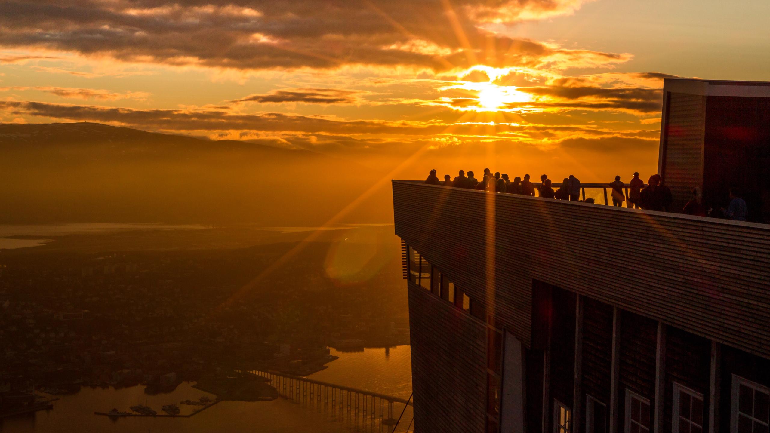 Folk står på takterrasen til Fjellheisen i Tromsø, mens midnatssolen farger himmelen oransje.