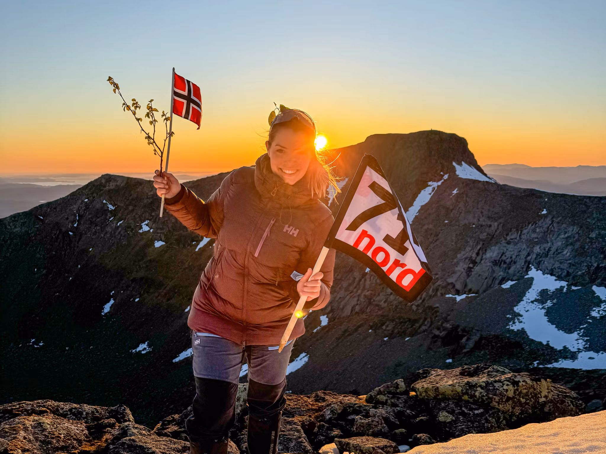 Woman waving the norwegian flag and a flag from the TV-production 71 grader nord in the sunset at a mountain in Rosendal, Hardanger