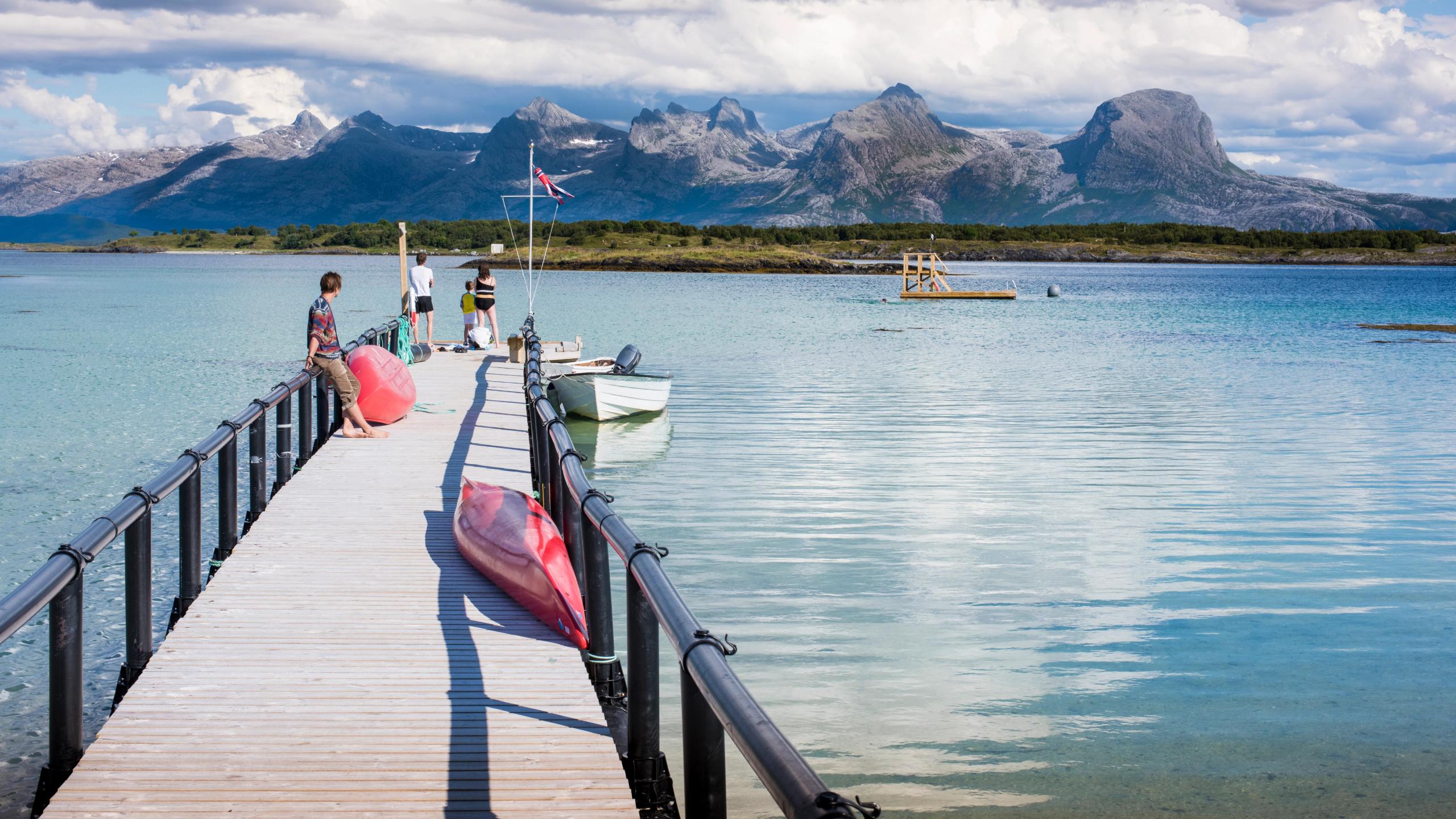 Folk står på en brygge på Helgelanskysten i Nordland. I bakgrunnen: fjellkjeden De syv søstre.