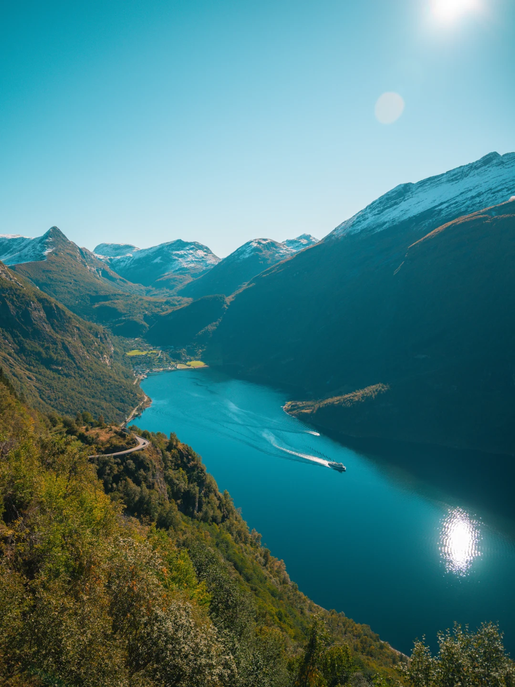 Overhead view from a hillside of a Norwegian fjord surrounded by snow-capped mountains, with a boat crossing the water.