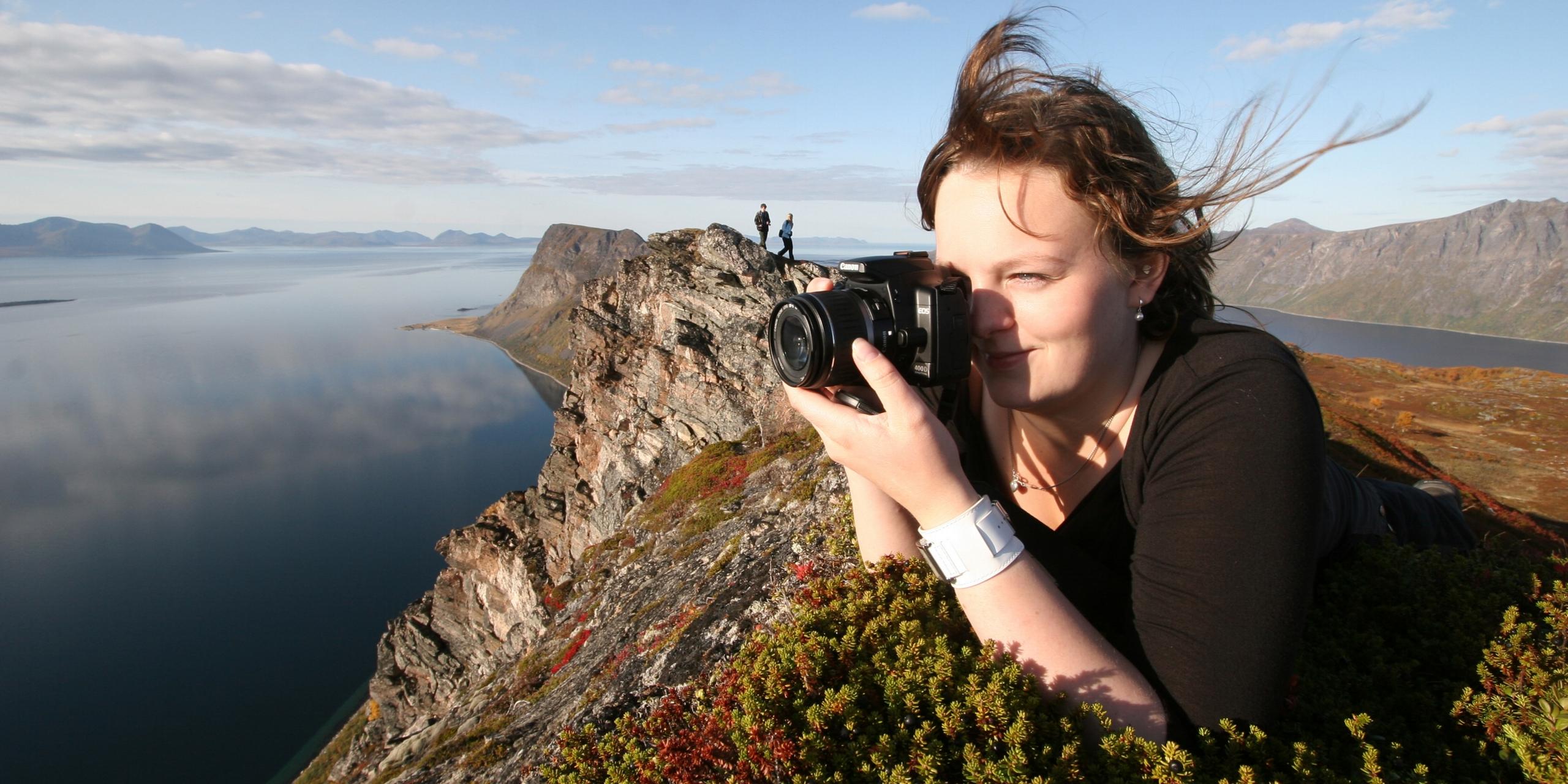 En jente ligger i lyngen på toppen av et fjell og tar bilde av horisonten