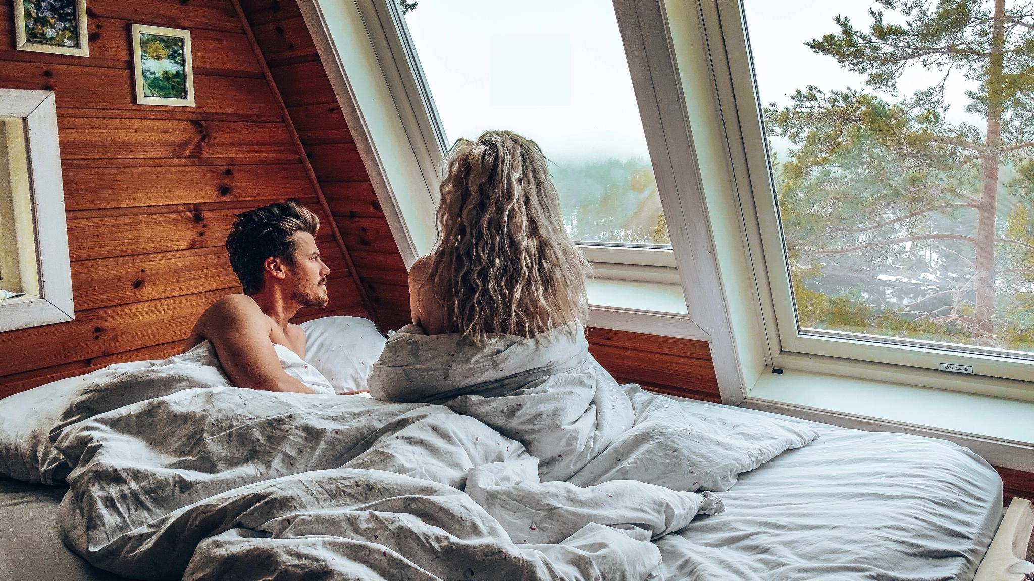 En dame og en mann sitter i sengen og ser ut av vinduet på Mikrohyttene i Kragerø i Telemark, Østlandet