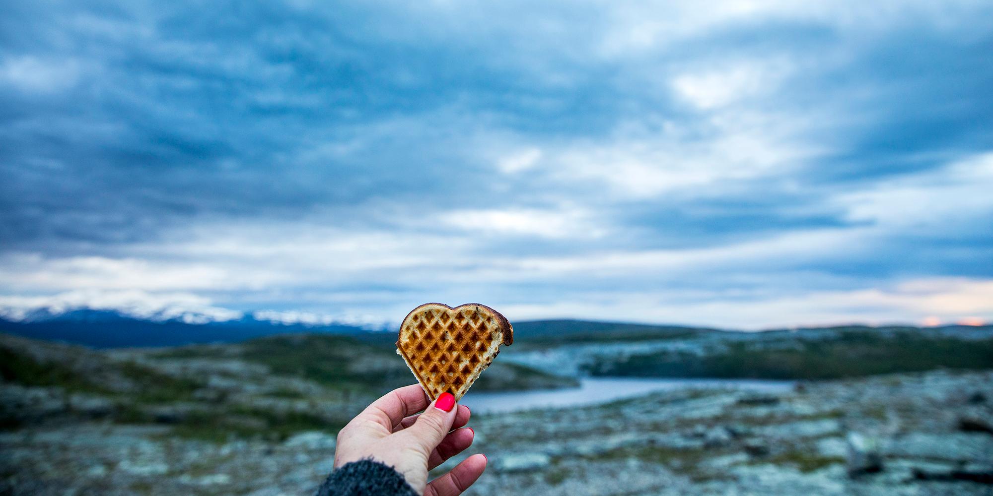 En hånd holder opp et vaffelhjerte med vakker natur som bakgrunn.