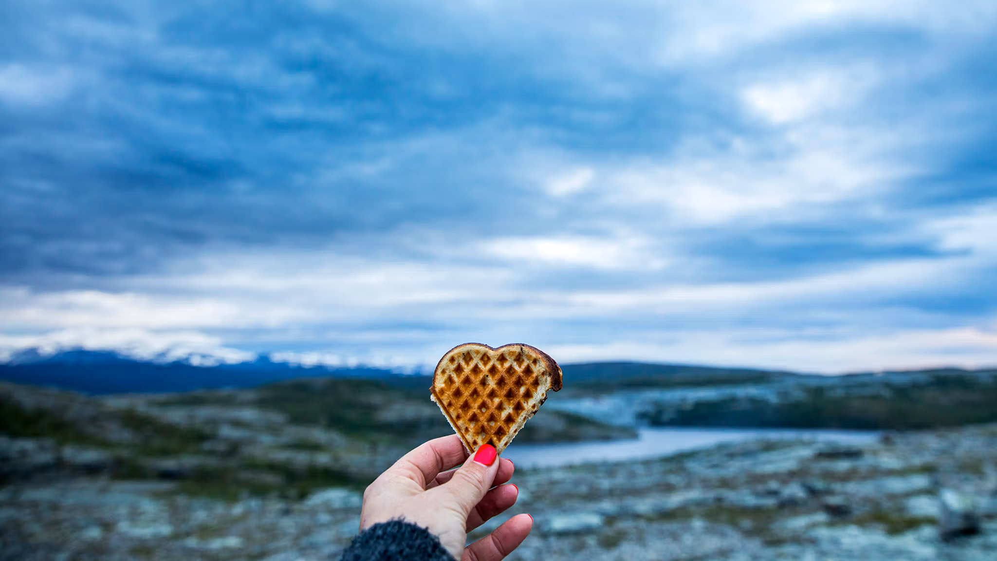 A hand holding up a heart-shaped waffle with beautiful scenery in the background