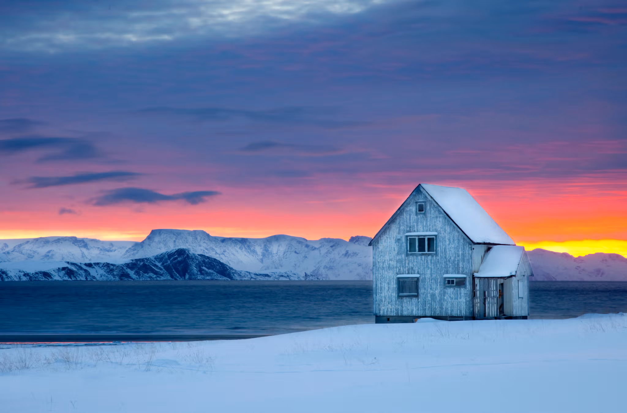 A wooden house next to the water on a snow-covered landscape with sunset colors in the background at Hasvik in Finnmark, Northern Norway